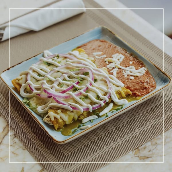 Enchiladas with green sauce, sour cream, red onions, and refried beans on a blue plate.