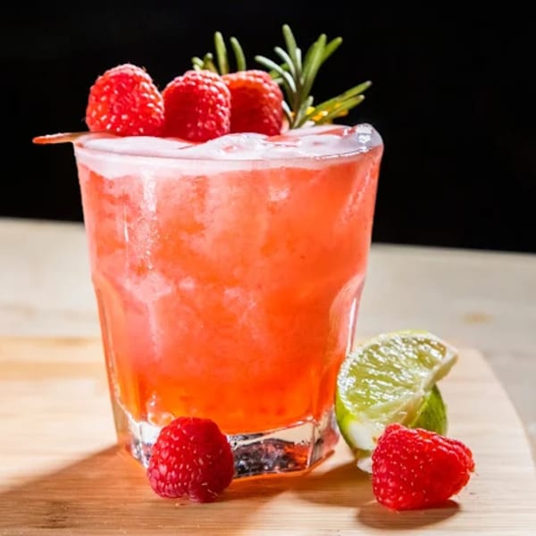 A vibrant red cocktail garnished with fresh raspberries and rosemary, served on a wooden board.