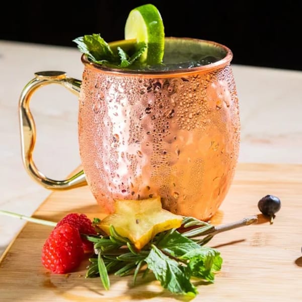A frosty copper mug with a drink, garnished with lime, mint, raspberry, starfruit, and rosemary.