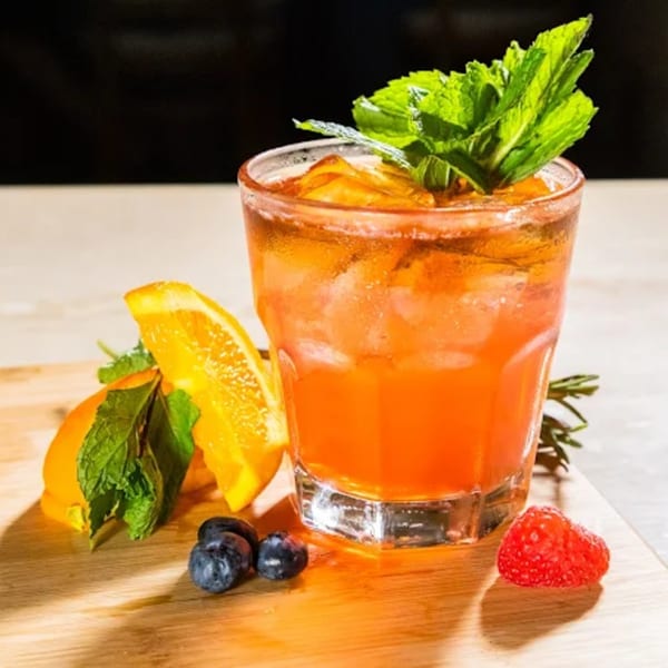 A vibrant orange cocktail with mint, orange slices, blueberries, and a raspberry on a wooden board.