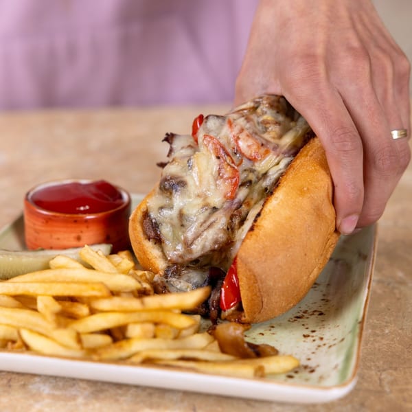 Hand holding a Philly cheesesteak sandwich with fries, pickles, and ketchup on a plate.