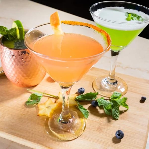Three vibrant cocktails on a wooden tray, garnished with mint, lime, grapefruit, starfruit, and blueberries.