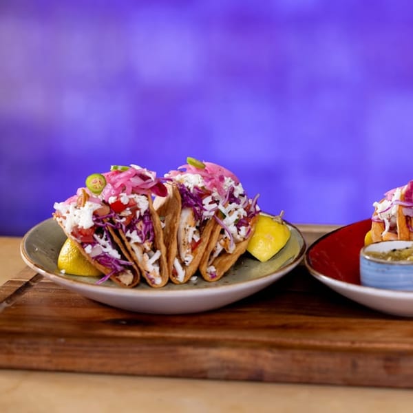 Three fish tacos with purple cabbage, white sauce, and pickled onions, garnished with lime wedges.
